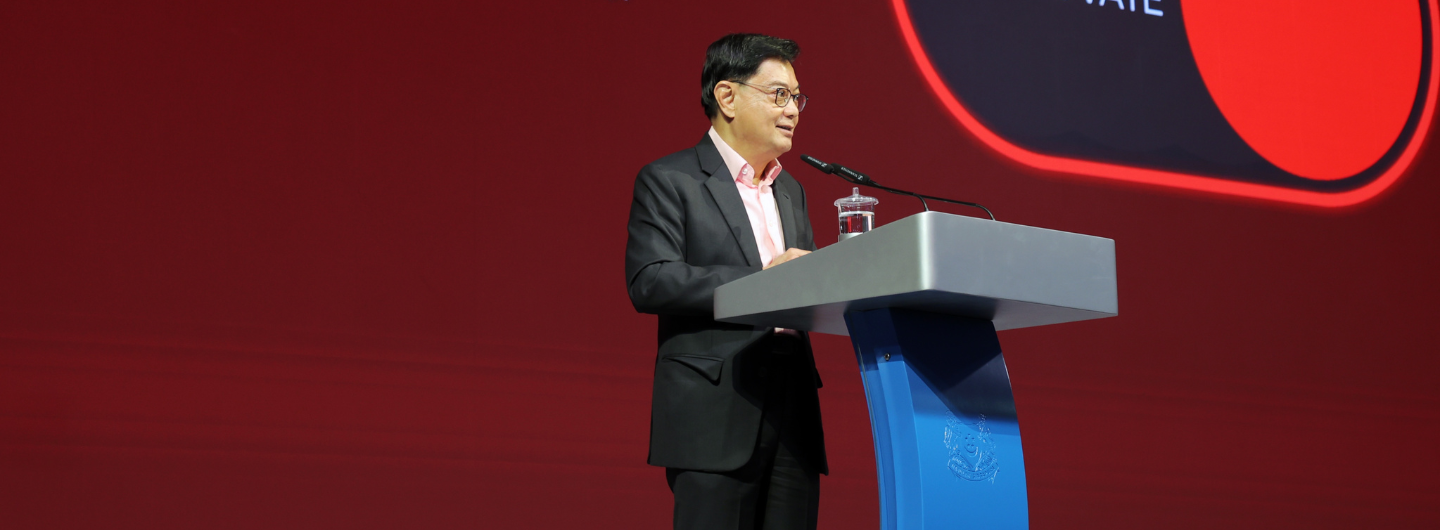 Man in suit at podium with Sennheiser mic, water, and Singapore crest, against a red backdrop.